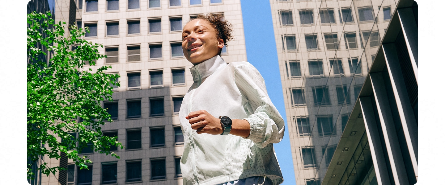 A woman in a workout outfit wearing a Galaxy Watch7 is seen walking on a city block surrounded by buildings.